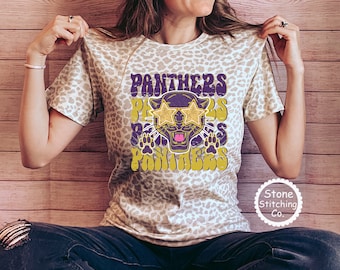 May include: A beige leopard print t-shirt with a purple and gold graphic of a panther wearing sunglasses and the text "Panthers" in a retro font.