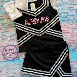 K&ouml;nnte beinhalten: Schwarzes Cheerleader-Outfit mit silbernen und wei&szlig;en Streifen. Das Oberteil hat einen V-Ausschnitt und der Rock ist kurz. Das Oberteil tr&auml;gt den Schriftzug "EAGLES" in maroonfarbener Schrift.