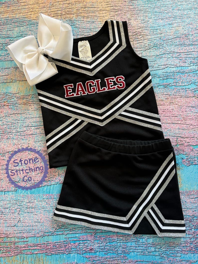 K&ouml;nnte beinhalten: Schwarzes Cheerleader-Outfit mit silbernen und wei&szlig;en Streifen. Das Oberteil hat einen V-Ausschnitt und der Rock ist kurz. Das Oberteil tr&auml;gt den Schriftzug "EAGLES" in maroonfarbener Schrift.