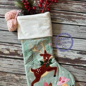 May include: A Christmas stocking with a light green background, featuring an embroidered reindeer with a wreath, mushrooms, and trees. The top is white, with pink pom-poms and a sprig of red berries. The text "Stone Stitching Co." is in a circle.