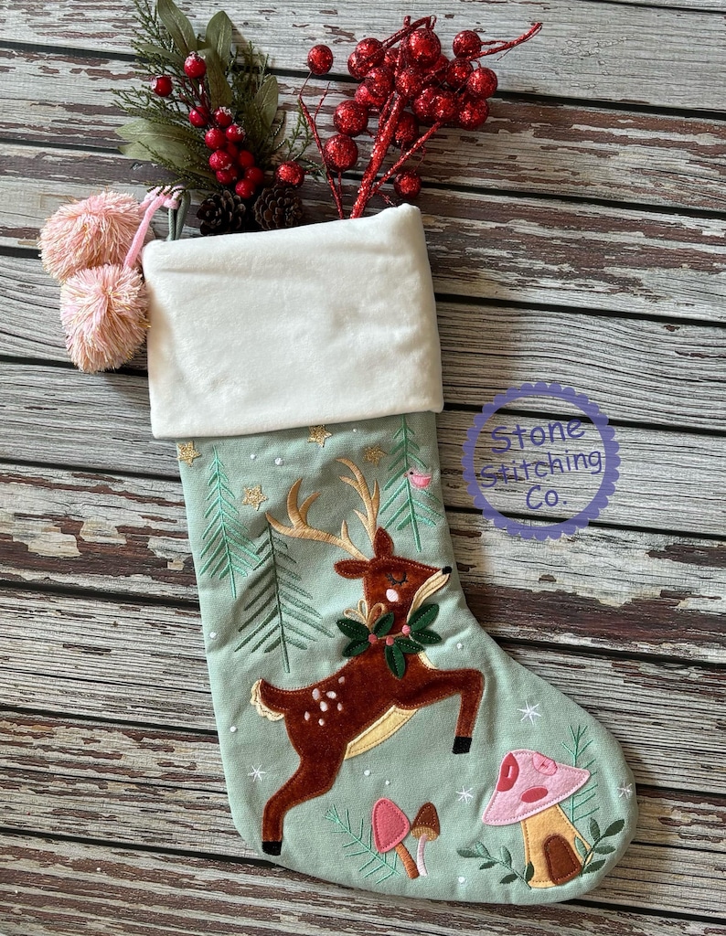 May include: A Christmas stocking with a light green background, featuring an embroidered reindeer with a wreath, mushrooms, and trees. The top is white, with pink pom-poms and a sprig of red berries. The text "Stone Stitching Co." is in a circle.