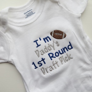 May include: White baby onesie with the text "I'm Daddy's 1st Round Draft Pick" embroidered in blue and gray, along with a brown football graphic. The onesie is labeled "Gerber" and is sized 0-3 months.