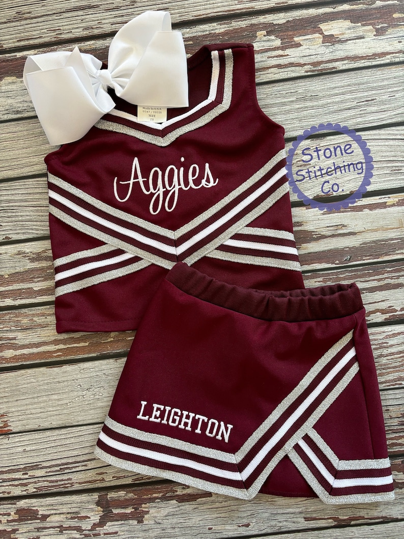 May include: Burgundy and silver cheerleading uniform with the word "Aggies" embroidered on the top and the name "Leighton" embroidered on the skirt.
