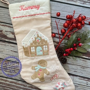 May include: A Christmas stocking with a gingerbread house and gingerbread man applique. The stocking is light pink with the name "Kammy" embroidered in pink. A candy cane and snowflake are also embroidered on the stocking.