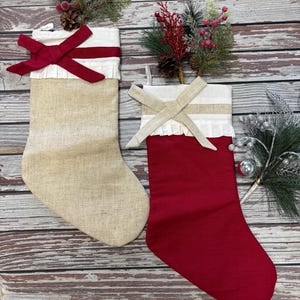 May include: Two Christmas stockings, one beige and one red, with white frilled trim and decorative bows. The beige stocking has a red bow, while the red stocking has a beige bow. The stockings are displayed on a wooden surface, with festive greenery.
