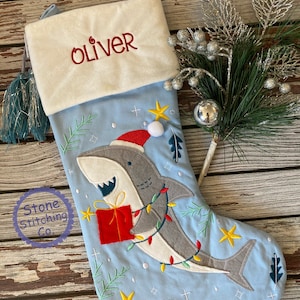 May include: Light blue Christmas stocking with a shark wearing a Santa hat and holding a red gift. The stocking has the name "Oliver" embroidered in red on the white cuff. Decorated with stars and Christmas lights.
