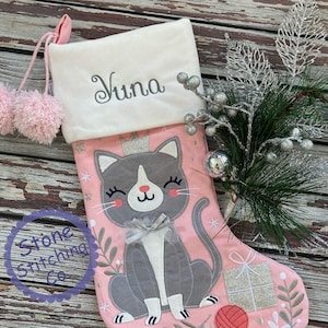 May include: Pink Christmas stocking with a gray cat design wearing a crown and a bow. The stocking has the name "Yuna" embroidered on the top. Decorative elements include a gift, yarn, and silver accents. The stocking is trimmed with a white cuff and pink pom-poms.