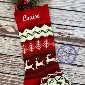 May include: Red and white knitted Christmas stocking with a green and white pattern featuring reindeer, trees, and snowflakes. The stocking is personalized with the name "Louise" in white embroidery.