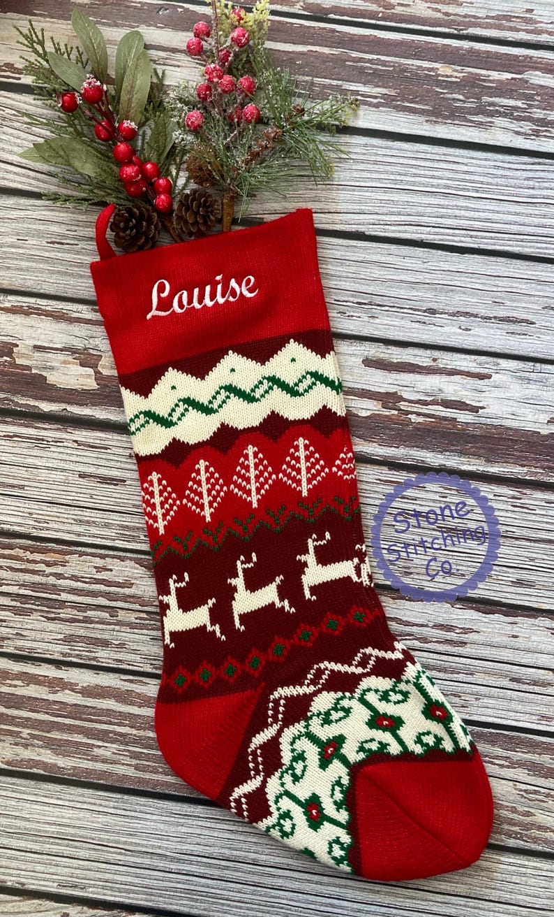 May include: Red and white knitted Christmas stocking with a green and white pattern featuring reindeer, trees, and snowflakes. The stocking is personalized with the name "Louise" in white embroidery.