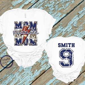 K&ouml;nnte beinhalten: Zwei wei&szlig;e T-Shirts. Ein Shirt hat den Text "MOM Football MOM" mit einem Fu&szlig;ball und einem Blitz-Motiv. Das andere Shirt hat den Text "SMITH 9" in Blau.