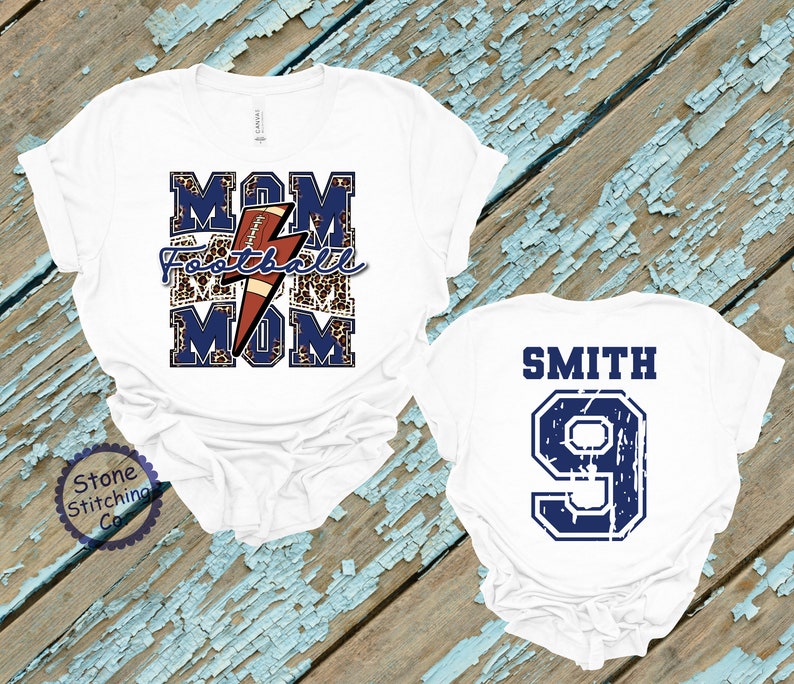K&ouml;nnte beinhalten: Zwei wei&szlig;e T-Shirts. Ein Shirt hat den Text "MOM Football MOM" mit einem Fu&szlig;ball und einem Blitz-Motiv. Das andere Shirt hat den Text "SMITH 9" in Blau.