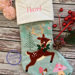 May include: A Christmas stocking with a light green background, featuring a brown deer with a green wreath, pink mushrooms, and the name "Hazel" embroidered in pink. The stocking has a white cuff and pink pom-poms.