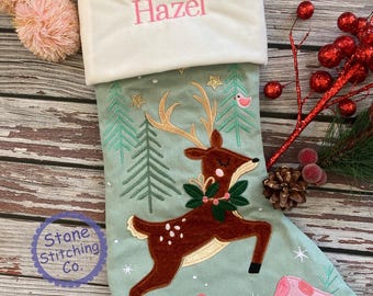 May include: A Christmas stocking with a light green background, featuring a brown deer with a green wreath, pink mushrooms, and the name "Hazel" embroidered in pink. The stocking has a white cuff and pink pom-poms.