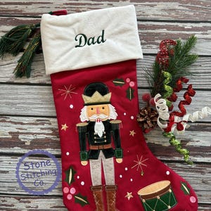 May include: Red Christmas stocking with a white cuff embroidered with the word "Dad" in green. The stocking features a nutcracker design, snowflakes, and a drum. The Stone Stitching Co. logo is visible.
