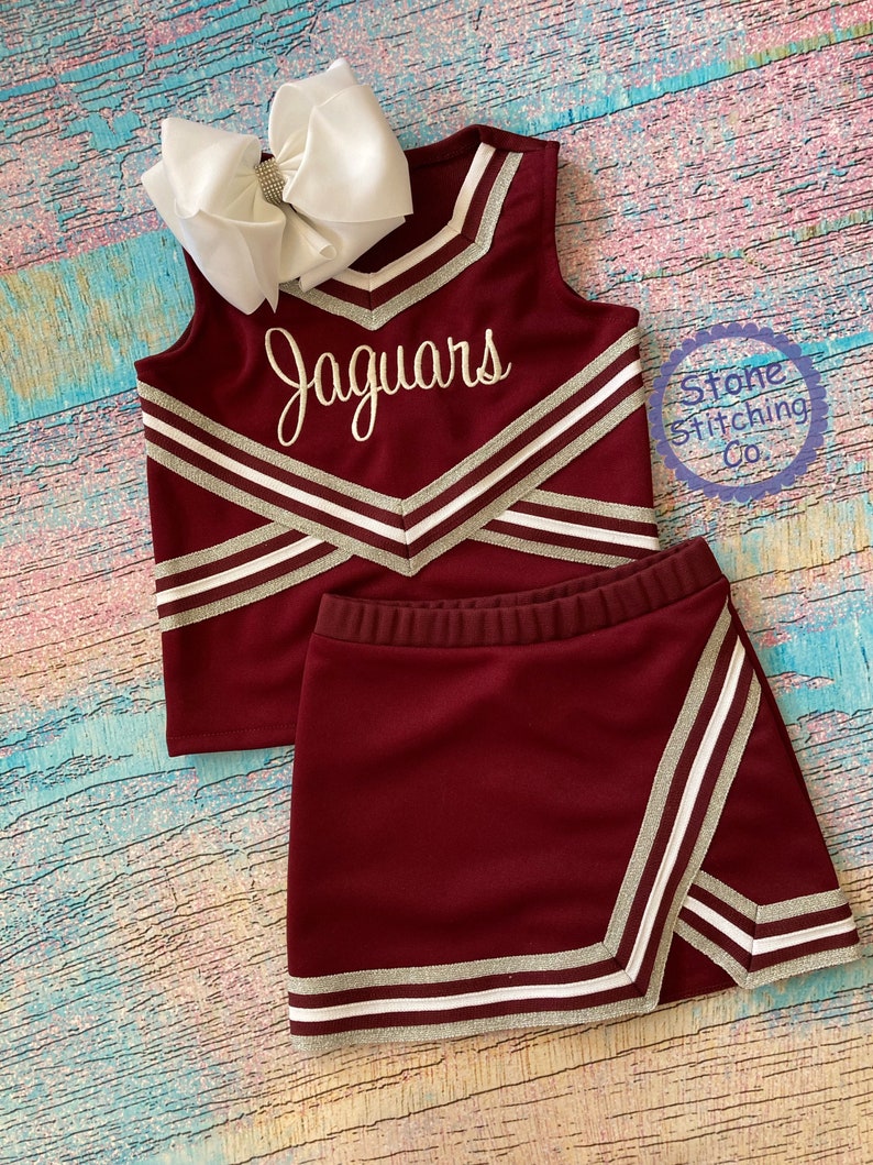 May include: Burgundy cheerleading uniform with silver and white stripes. The top has the word "Jaguars" embroidered on the front. The skirt is a mini skirt with a side panel.