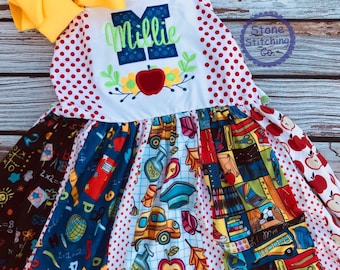 May include: A white sleeveless dress with a red polka dot pattern. The dress has a multi-colored patchwork skirt with school-themed patterns. The top of the dress has a blue and green embroidered letter M and the name Millie.