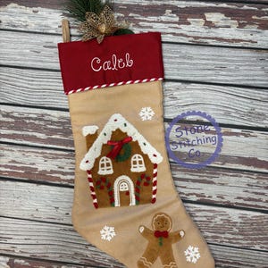May include: A tan Christmas stocking with a red cuff embroidered with the name "Caleb". The stocking features a gingerbread house and gingerbread man design with white snowflakes. A sprig of greenery and gold bow adorn the top.