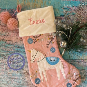 May include: A pink Christmas stocking featuring a white unicorn design, blue baubles, and the name "Lexie" embroidered in pink. The stocking has a cream cuff and pink pom-poms. The Stone Stitching Co. logo is visible.