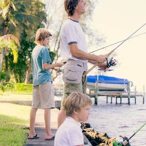 Pode incluir: Um pai e dois filhos pescando de um cais. O pai está segurando uma vara de pesca e olhando para o céu. O filho mais velho está segurando uma vara de pesca e olhando para a água. O filho mais novo está sentado em uma rocha e segurando uma vara de pesca.