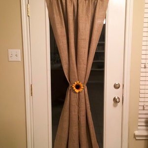 May include: A brown burlap door curtain with a sunflower tied at the center. The curtain is hung on a white door with a glass panel.