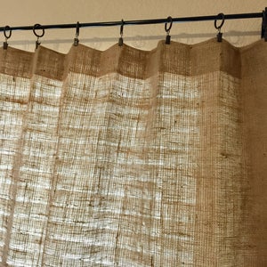 May include: A pair of brown burlap curtains hanging from a black metal curtain rod. The curtains are made of a woven fabric with a rough texture.