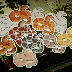 Puede incluir: Una colección de pegatinas de serpientes en varios colores y patrones, incluyendo naranja, marrón, gris y amarillo. Las pegatinas presentan una ilustración de estilo caricatura de una serpiente con la lengua fuera.