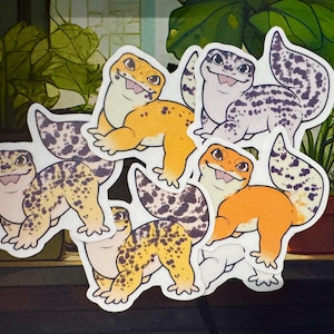 Puede incluir: Una colección de pegatinas de gecko coloridas. Las pegatinas presentan geckos de dibujos animados en varias poses y colores, incluyendo naranja, amarillo y morado. Los geckos tienen patrones de manchas y están sonriendo. El fondo incluye plantas en macetas.