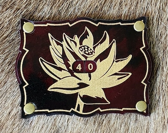 Può includere: Una scatola in pelle bordeaux con un design a fiore di loto dorato e un bottone in metallo dorato su ogni lato. La scatola ha un numero "40" in metallo dorato sul davanti.
