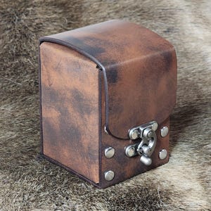 Peut inclure: Une boîte en cuir marron avec un fermoir et des rivets en métal. La boîte a un aspect vieilli et est parfaite pour ranger de petits objets.