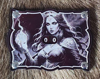 Può includere: Un'immagine in bianco e nero di una donna con lunghi capelli scuri che indossa una corona e un abito fluente. Tiene un globo luminoso nella mano. L'immagine è su uno sfondo nero con un bordo argentato e rivetti argentati. L'immagine ha un contatore con i numeri 00.