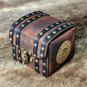 May include: A small, square, brown leather chest with black straps and brass rivets. The chest has a brass clasp and a circular emblem with a skull design. The chest is designed to look like a treasure chest.
