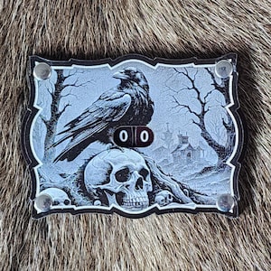 May include: A rectangular, black and white decorative piece featuring a raven perched atop a skull. The artwork includes a spooky landscape with bare trees and a haunted house. The piece has silver rivets and displays the numbers "00".