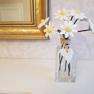May include: A small glass vase with a white heart charm tied around the neck. The vase is filled with a bouquet of white ceramic daisies with yellow centres.