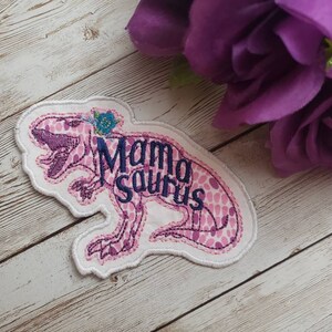 May include: Pink and white embroidered iron-on patch with a pink dinosaur silhouette and the text "Mama Saurus".