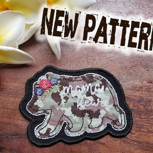 May include: A brown and green camouflage fabric bear applique with a black outline. The bear has a floral crown with red and blue flowers and the text "mama bear" embroidered in white.