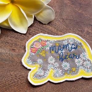 Peut inclure: Une applique en tissu gris en forme d'ours avec une bordure jaune. L'ours est décoré de marguerites blanches et des mots "mama bear" en broderie bleue.