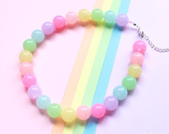 Può includere: Una collana di perline arcobaleno pastello con una catena d'argento. La collana presenta perline rotonde nei toni del rosa, giallo, verde, blu e viola. Lo sfondo è una sfumatura di colori pastello, con il testo "let's go fairy!"