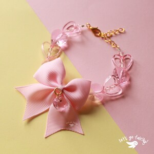 May include: Pink heart-shaped charm bracelet with a pink bow and a gold chain. The bracelet has the text "let's go fairly!" on it.