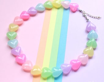 Può includere: Una collana di perline a forma di cuore dai colori pastello: rosa, viola, giallo, verde e blu. La collana è su uno sfondo rosa con una striscia arcobaleno. Il testo "let's go fairy!" è nell'angolo in basso a destra.