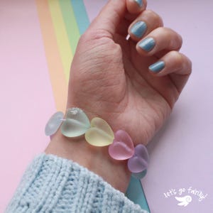 May include: A bracelet made of heart-shaped beads in pastel colours: light blue, yellow, pink, and purple. The text "let's go fairy!" is visible. The bracelet is worn on a wrist.