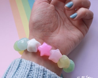 Può includere: Un braccialetto a forma di stella con perline bianche, rosa e gialle. Il braccialetto è indossato su un polso, con un maglione azzurro chiaro sullo sfondo. Il testo "let's go fairly!" è visibile.