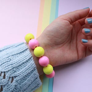 Könnte beinhalten: Ein Armband mit abwechselnd gelben und rosa runden Perlen. Das Armband wird an einem Handgelenk mit einem hellblauen Strickärmel getragen. Der Hintergrund ist eine Pastellfarbpalette aus Rosa, Gelb und Blau.