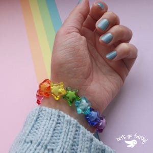 May include: A colourful star-shaped bracelet with beads in rainbow hues. The bracelet is worn on a wrist with blue nail polish. The background includes a rainbow and the text "let's go fairy!". The bracelet is made of translucent beads in shades of red, orange, yellow, green, blue, and purple.