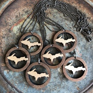 May include: Six wooden necklaces with a cutout of the Upper Peninsula of Michigan. The necklaces are on a rustic metal surface.