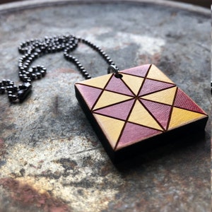 Può includere: Una collana con un ciondolo quadrato in legno con un motivo geometrico di triangoli gialli e bordeaux. Il ciondolo è attaccato a una catena nera.
