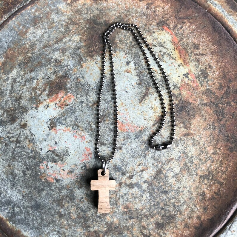 May include: A wooden cross pendant on a silver ball chain necklace. The necklace is laying on a rustic metal surface.