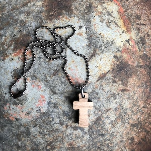 May include: A wooden cross pendant on a silver ball chain necklace. The necklace is lying on a textured, rusty metal surface.