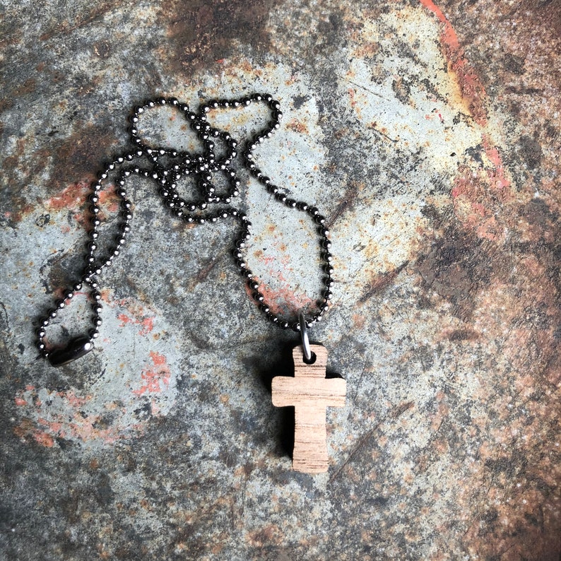 May include: A wooden cross pendant on a silver ball chain necklace. The necklace is lying on a textured, rusty metal surface.