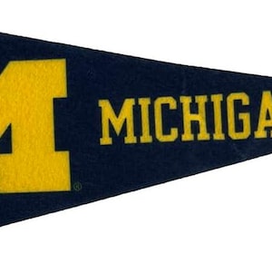 Puede incluir: Un banderín azul marino con una "M" amarilla y la palabra "MICHIGAN" en amarillo. Tiene una franja vertical amarilla en el lado izquierdo. El banderín tiene forma triangular.
