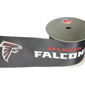May include: A spool of black ribbon with the Atlanta Falcons logo and team name in white and red. The NFL logo is on the spool. The ribbon is on a white background.